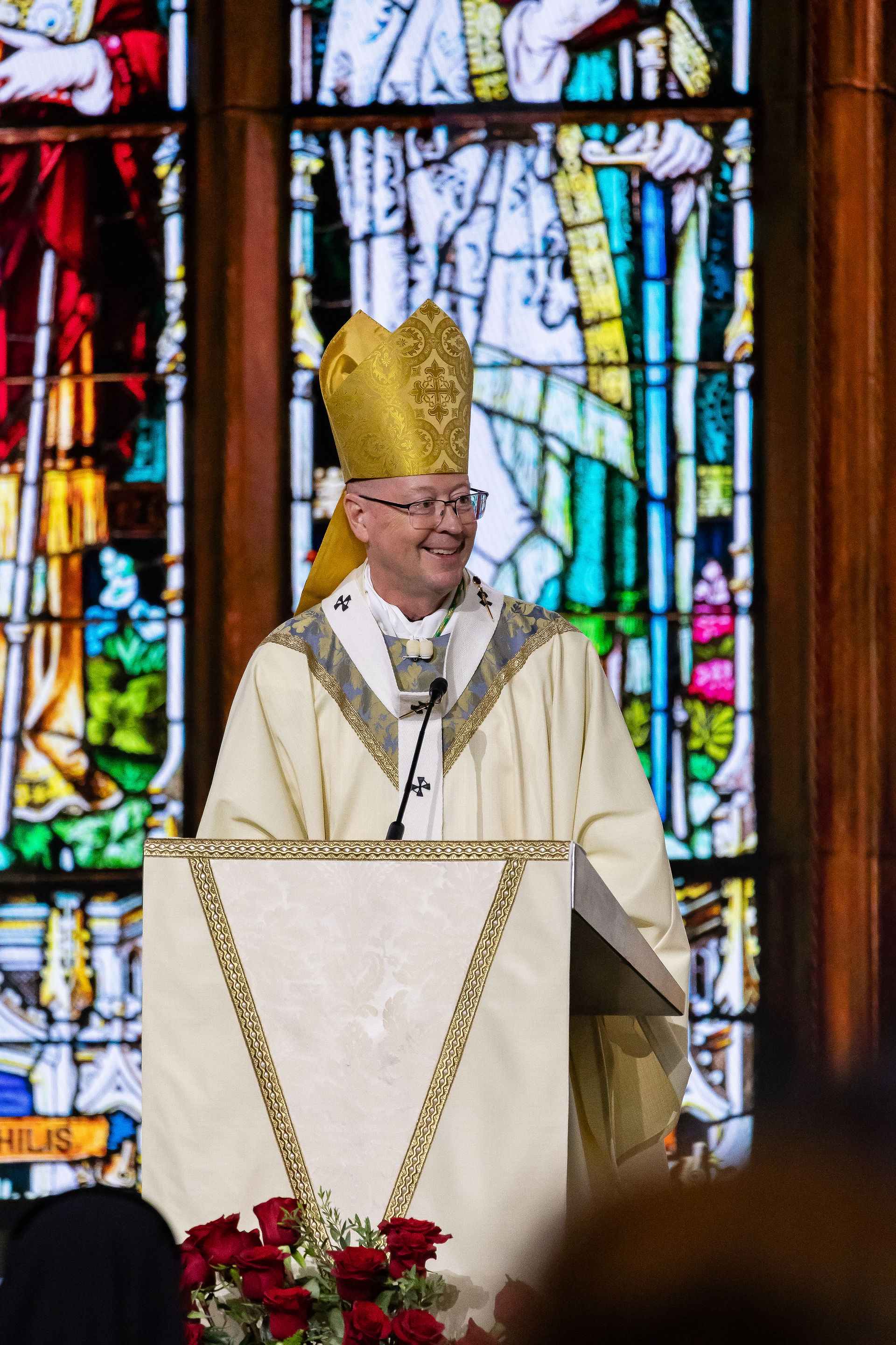 Smiling bishop in gold vestments and mitre speaks at a church lectern before colorful stained glass.