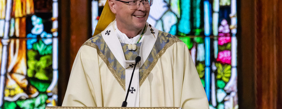 Smiling bishop in gold vestments and mitre speaks at a church lectern before colorful stained glass.