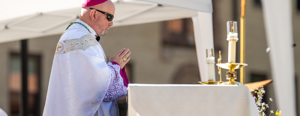 Catholic bishop in white and purple vestments prays at an outdoor altar with candles under a canopy, in a solemn ceremony