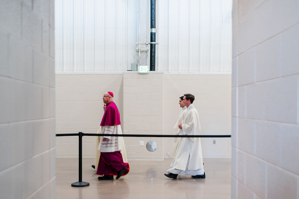 Three clergy members walk through a bright hallway. One wears maroon robes, the others in white. An exit sign is visible above.
