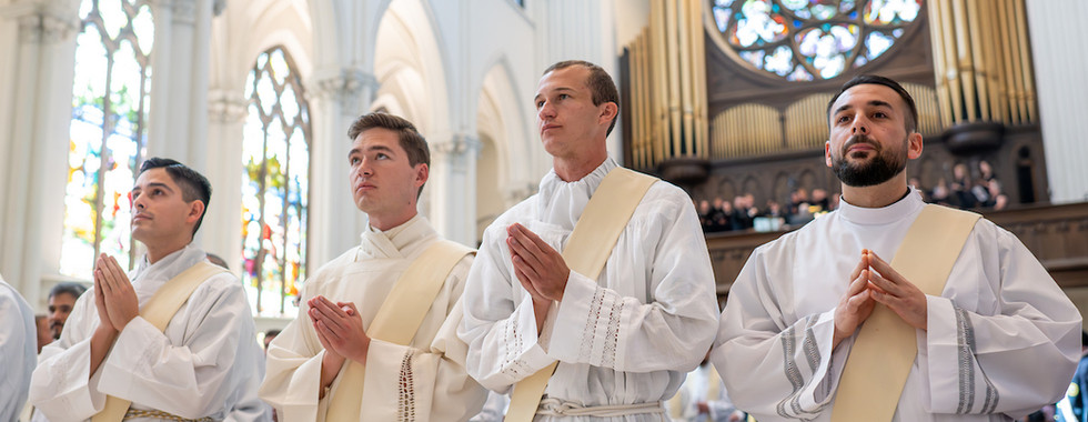 Four men in white robes and sashes pray inside a cathedral with stained glass and organ pipes in the background. They appear solemn and focused.
