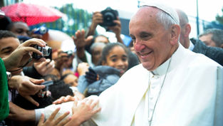 A religious figure in white attire smiles, greeting a crowd under umbrellas. Cameras capture the moment, creating a joyful atmosphere.