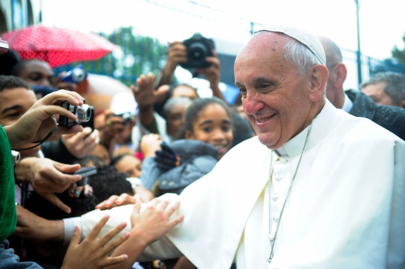 A religious figure in white attire smiles, greeting a crowd under umbrellas. Cameras capture the moment, creating a joyful atmosphere.