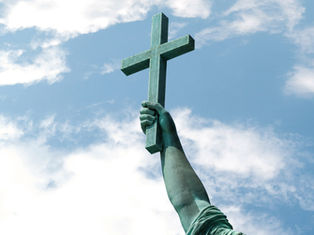 Brazo de estatua verde sosteniendo una cruz de madera contra un cielo azul con nubes, evocando solemnidad.