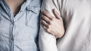 Person in denim shirt stands next to another in white, with an arm linked and hand displaying rings, creating an intimate, close atmosphere.