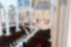 Wide view of a cathedral interior with stained glass windows. Clergy in robes perform a ceremony at the altar, while people observe from pews.