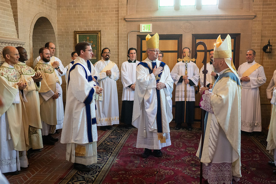 A group of clergy in ornate robes and miters stand in a church interior, engaged in conversation. The mood is formal and solemn.