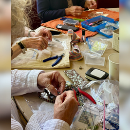 Hands make beaded jewelry at a craft table with pliers, beads, trays, and colorful tools on towels.