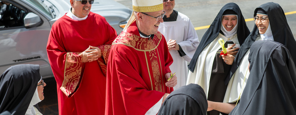 Clergy in red robes and nuns with veils gather on a street, smiling and interacting. A priest wears a mitre. Bright and sunny day.