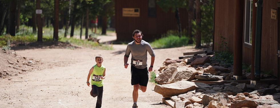 Father and son jogging on a dirt path in a wooded area, wearing race numbers. They're smiling, with cabins and trees in the background.