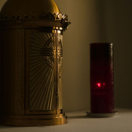 Golden tabernacle with engraved cross beside a glowing red candle in a dimly lit setting, creating a serene and reverent atmosphere.