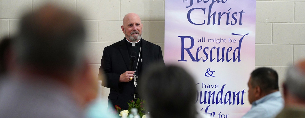 Bald man in black clergy attire speaks at an event, standing by a banner with Christian text. Attendees sit in foreground. Neutral mood.