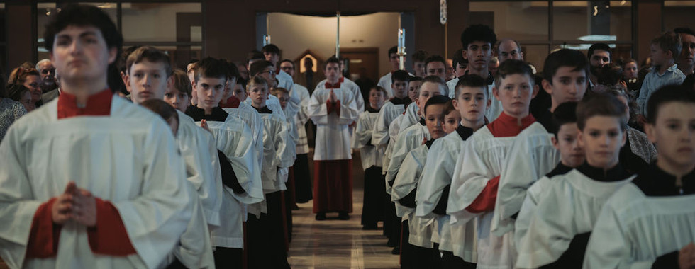 Dozens of altar servers line up down the center aisle of a Catholic church. One holds a large processional cross in the center. They face the altar and the back of the church is visible.