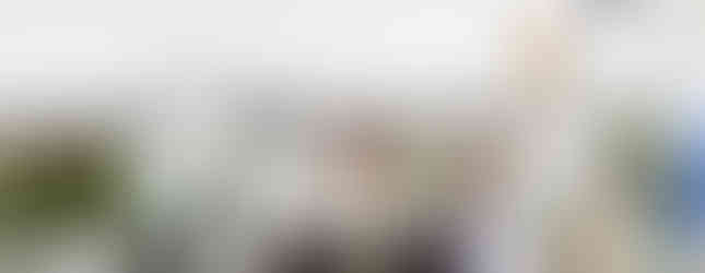 Priests in white robes conduct a religious ceremony under a white canopy. One holds a chalice, others kneel. Trees and a cloudy sky in the background.