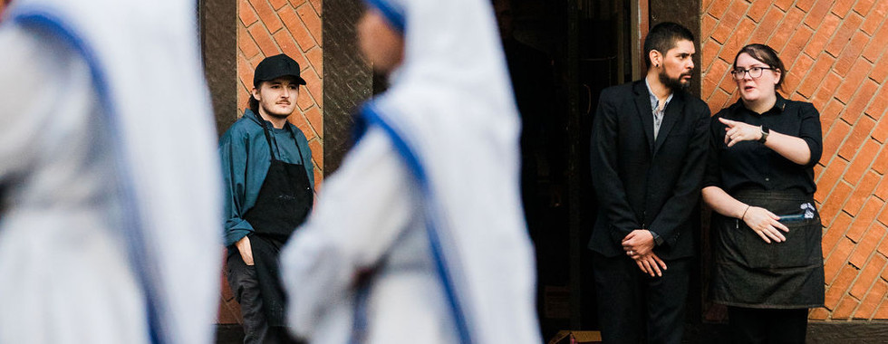 Blurred nuns walk past a brick building with an Associates Entrance sign, while two staff stand talking at the doorway.