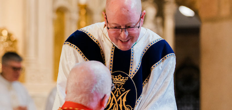 A priest in white and blue robes leans towards a seated man in red, inside a church with ornate architecture. Warm, solemn atmosphere.