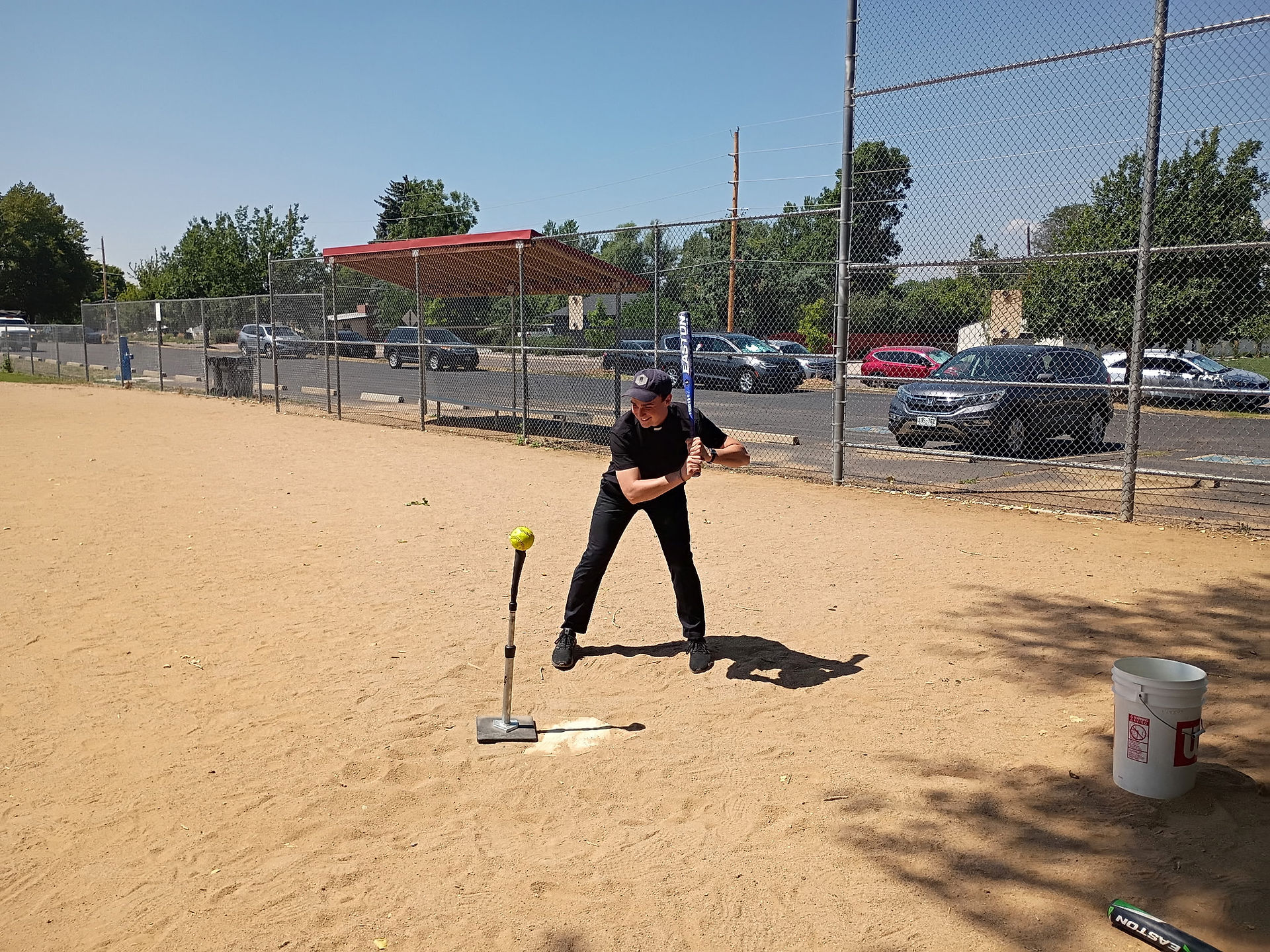 Man in black at a softball tee on a dusty field, ready to hit a yellow ball; chain-link fence, cars, and blue sky behind him