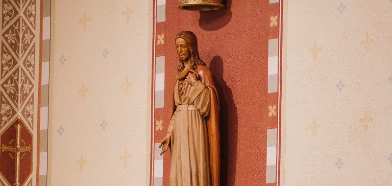 Wooden statue of Jesus in a church niche with red paneling, gold crown above, and patterned cream walls.