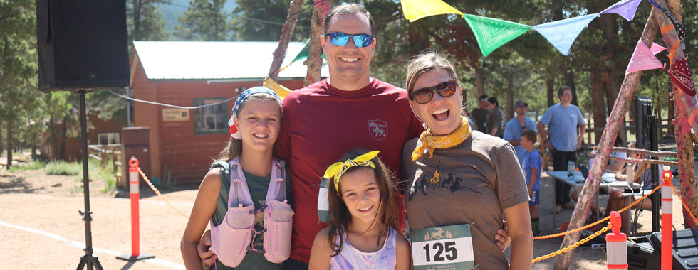 Smiling family of four posing outdoors at a mountain event with colorful bunting; one woman wears bib 125.