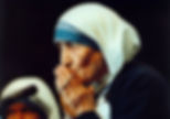 Mother Teresa in white-and-blue habit prays with clasped hands near a microphone against a dark background.