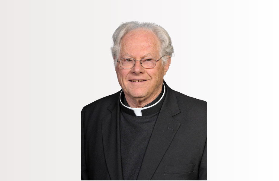 Portrait of an elderly priest in black clerical attire and glasses, smiling against a plain white background.