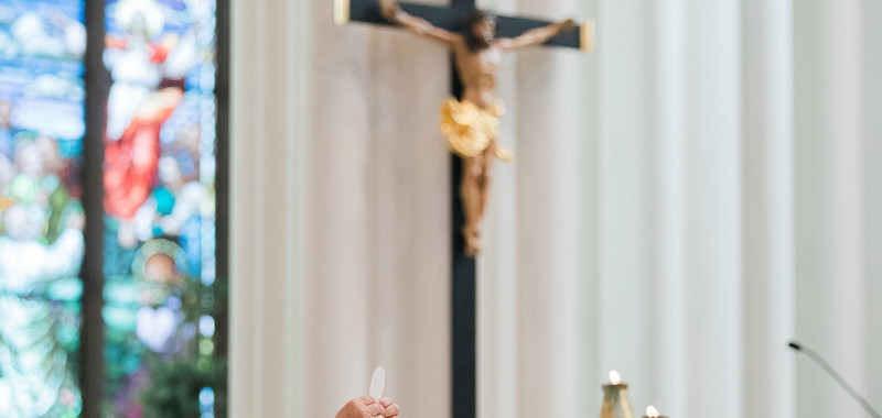 Priest in white robe raises the Eucharist during Mass in a church. Stained glass and candles are visible, with a crucifix in the background.
