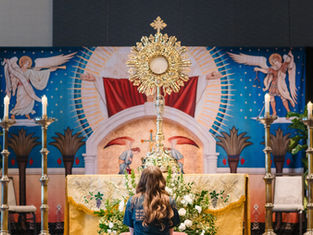 A person kneels facing a ornate monstrance on an altar with candles. Religious mural backdrop features angels, rich colors, and patterns.