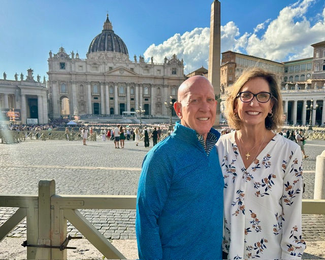 Smiling couple in front of St. Peter's Basilica, Rome, on a sunny day. Blue sky, ornate architecture, and cobblestone plaza in the background.