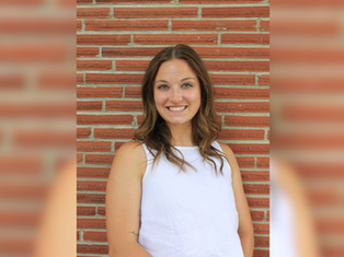 Smiling woman in a white top stands against a brick wall. She's looking at the camera, creating a cheerful mood.