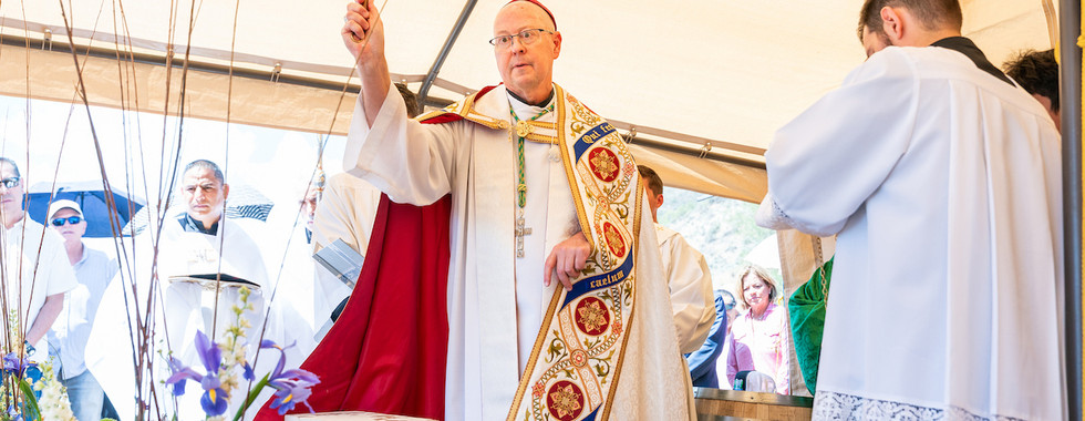 Bishop in white and red vestments raises a staff at an outdoor Mass under a tent, with flowers and worshippers nearby