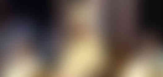 A bishop in ornate robes and mitre sits on a grand wooden chair, flanked by two priests in a ceremonial setting, with onlookers blurred in the background.