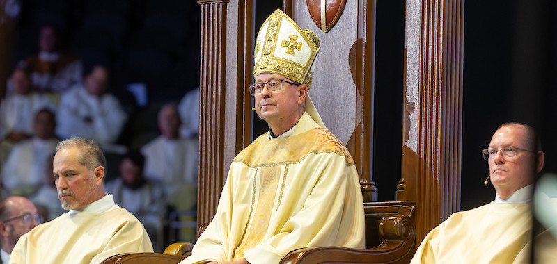 A bishop in ornate robes and mitre sits on a grand wooden chair, flanked by two priests in a ceremonial setting, with onlookers blurred in the background.