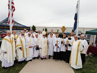 Clergy and altar servers in white and gold robes pose outdoors by a flagpole. "Rocky Mtn Regional VA Medical Center" visible on a canopy.