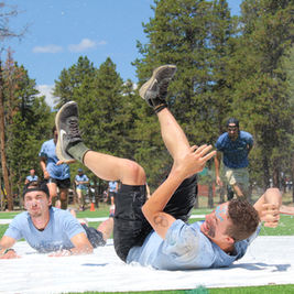 People tumble on a wet outdoor slip-n-slide, splashing on grass under pine trees in a sunny park.