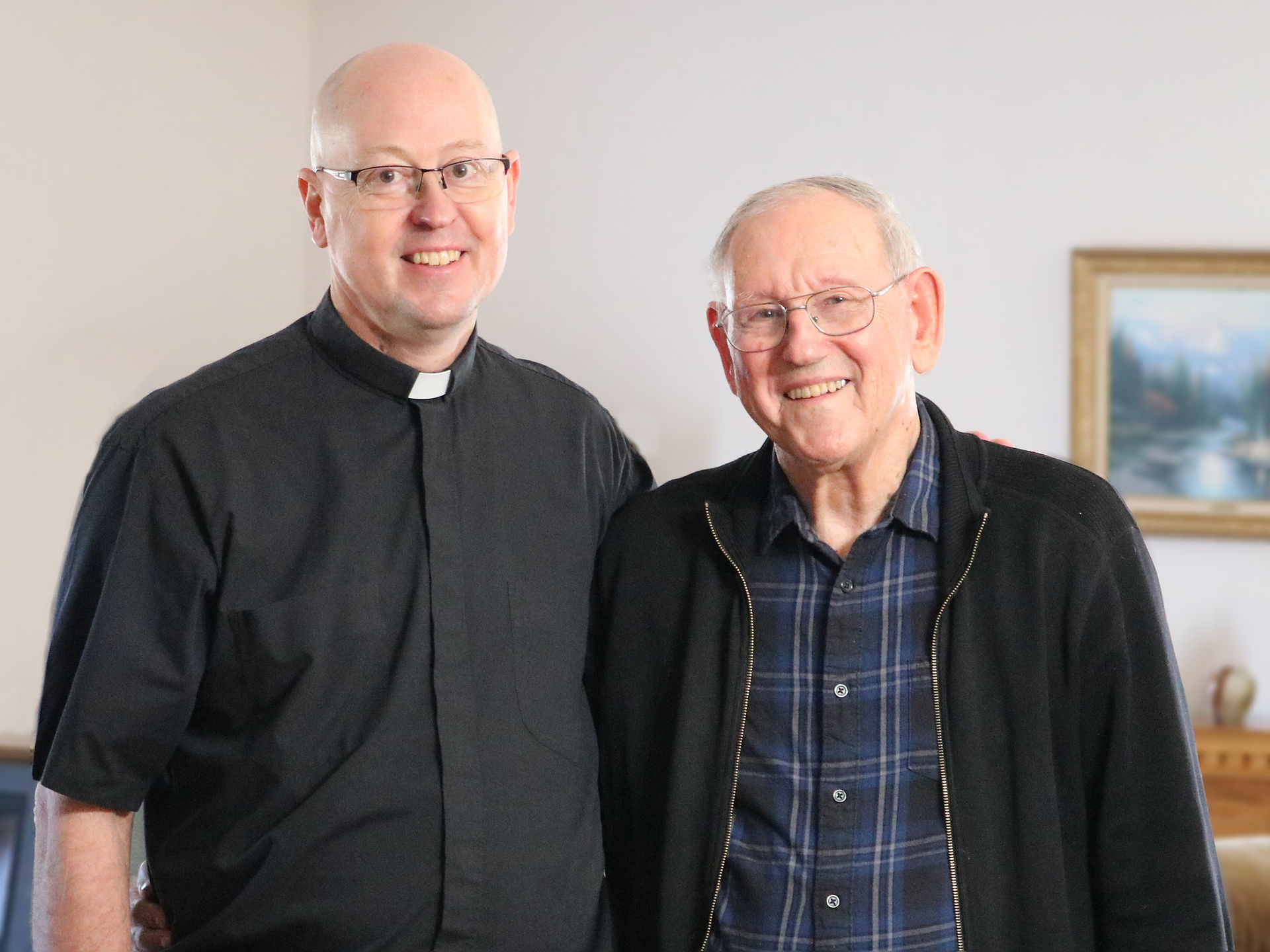Two smiling men stand side by side indoors; one in a priest collar, the other in plaid. A framed landscape painting is visible in the background.