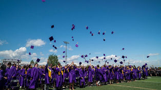 Graduates in purple gowns toss caps into a bright blue sky on a football field, celebrating at an outdoor ceremony.