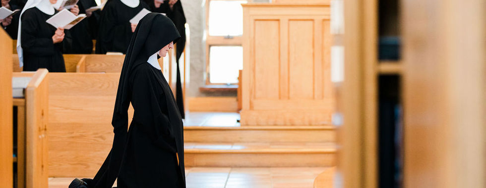A religious sister kneels in prayer before the altar as fellow sisters sing in the background during the profession liturgy.