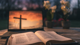 Open Bible on a wooden table in front of a laptop showing a cross at sunset, with blurred flowers and a warm, reverent mood