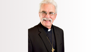 Elderly man with glasses and mustache smiling, wearing a black suit and clerical collar with a cross necklace, on a plain white background.
