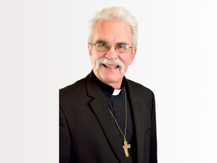 Elderly man with glasses and mustache smiling, wearing a black suit and clerical collar with a cross necklace, on a plain white background.
