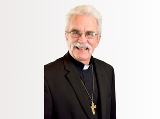 Elderly man with glasses and mustache smiling, wearing a black suit and clerical collar with a cross necklace, on a plain white background.