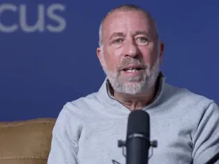 Man with a beard speaking into a microphone against a blue background with partial "CUS" text visible. He wears a gray sweater, seated on a brown couch.