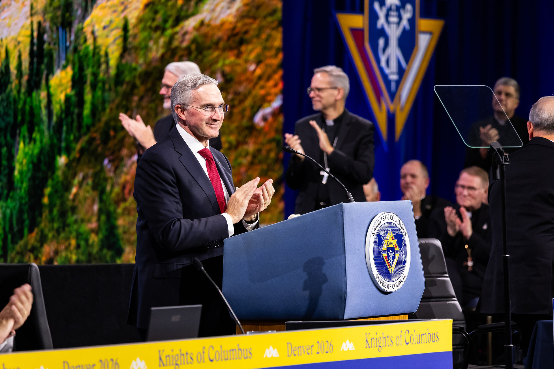 Man in suit and red tie speaks at Knights of Columbus podium as clergy and attendees applaud onstage, Denver 2026 banner.