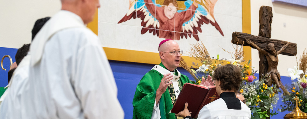 Catholic bishop in green vestments blesses a book during Mass, with altar servers, crucifix, and flowers in a church.