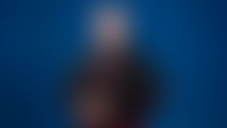 Un hombre con atuendo religioso negro y detalles rojos, sonriendo frente a un fondo azul. Lleva un anillo y collar. Ambiente formal.