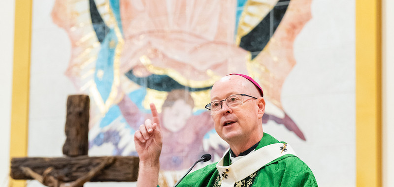 Bishop in green vestments speaks at a church lectern, raising one finger, with a crucifix and large Virgin Mary image behind.
