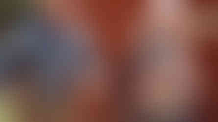 Older man and woman joyfully jog near a red cabin in a sunny outdoor setting. The man wears a cowboy hat and holds a camera.