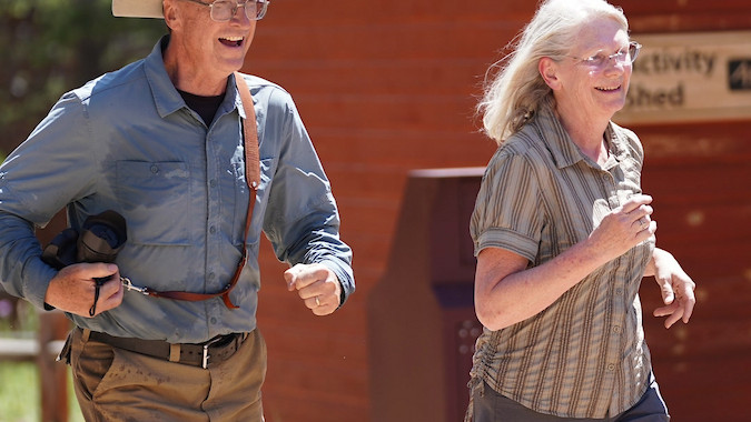 Older man and woman joyfully jog near a red cabin in a sunny outdoor setting. The man wears a cowboy hat and holds a camera.