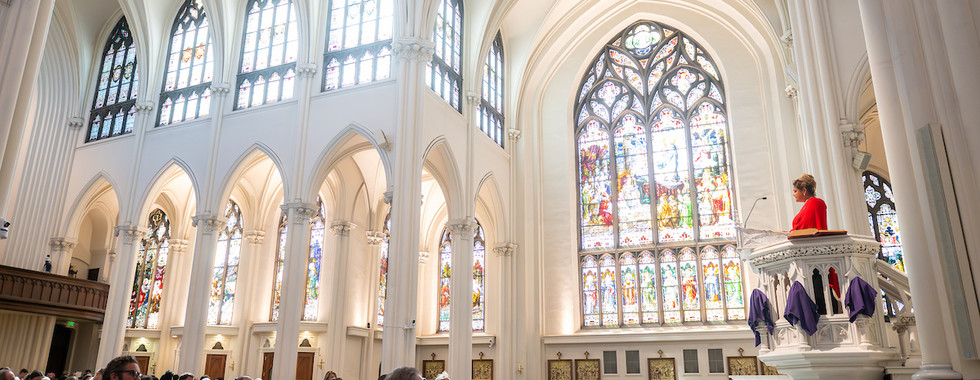 Wide view of a cathedral interior with stained glass windows as a lector reads from the ambo during the liturgy.