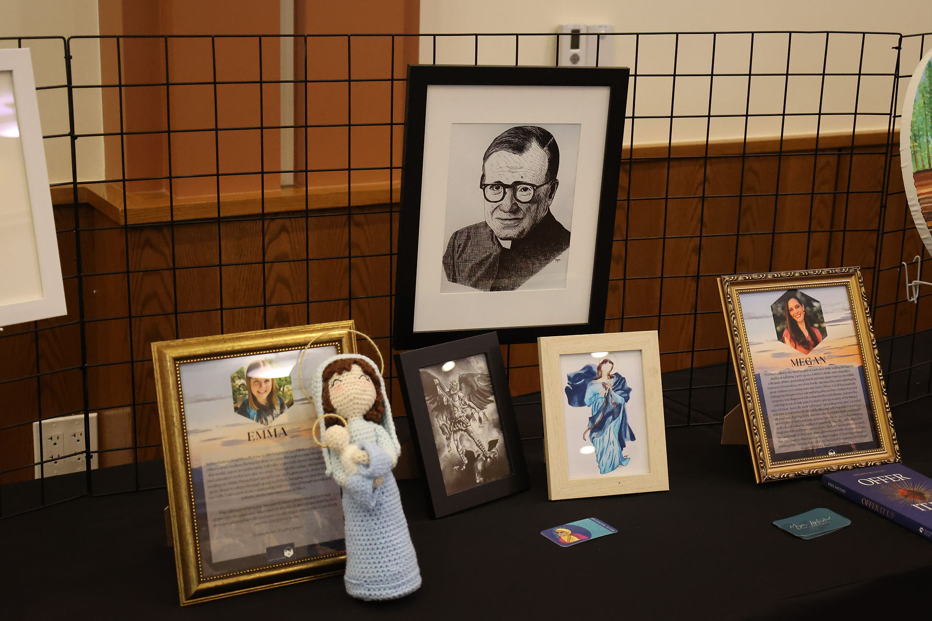 Display table with framed portraits, religious artwork, Marian imagery and handmade devotional items at a Catholic fundraising event.